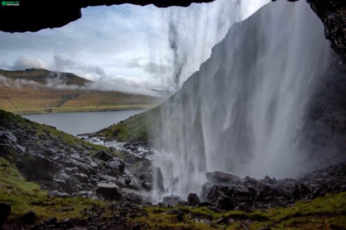 View from behind a waterfall in the Faroe Islands, looking out through the curtain of falling water towards a fjord lake and misty mountains beyond