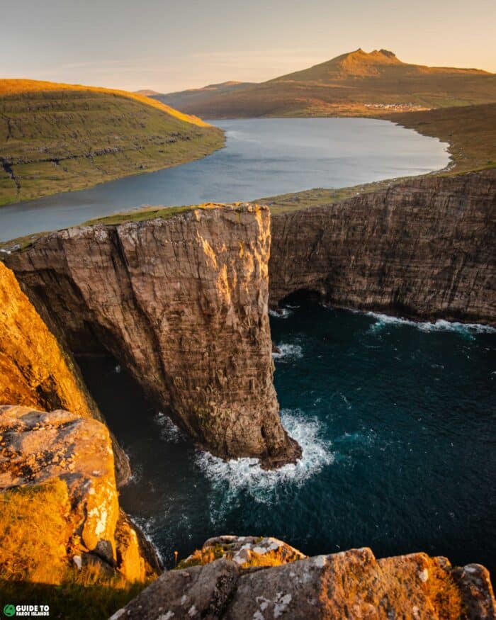 Lake Leitisvatn appearing to float above the North Atlantic Ocean, separated by towering vertical cliffs with crashing waves below, viewed from above at golden hour on Vágar Island, Faroe Islands