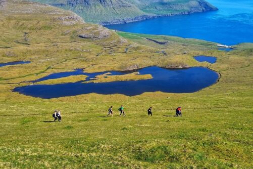 Group of hikers ascending a green mountain slope on Streymoy Island, Faroe Islands, with a dark blue highland lake and vivid fjord coastline visible below on a sunny summer day.