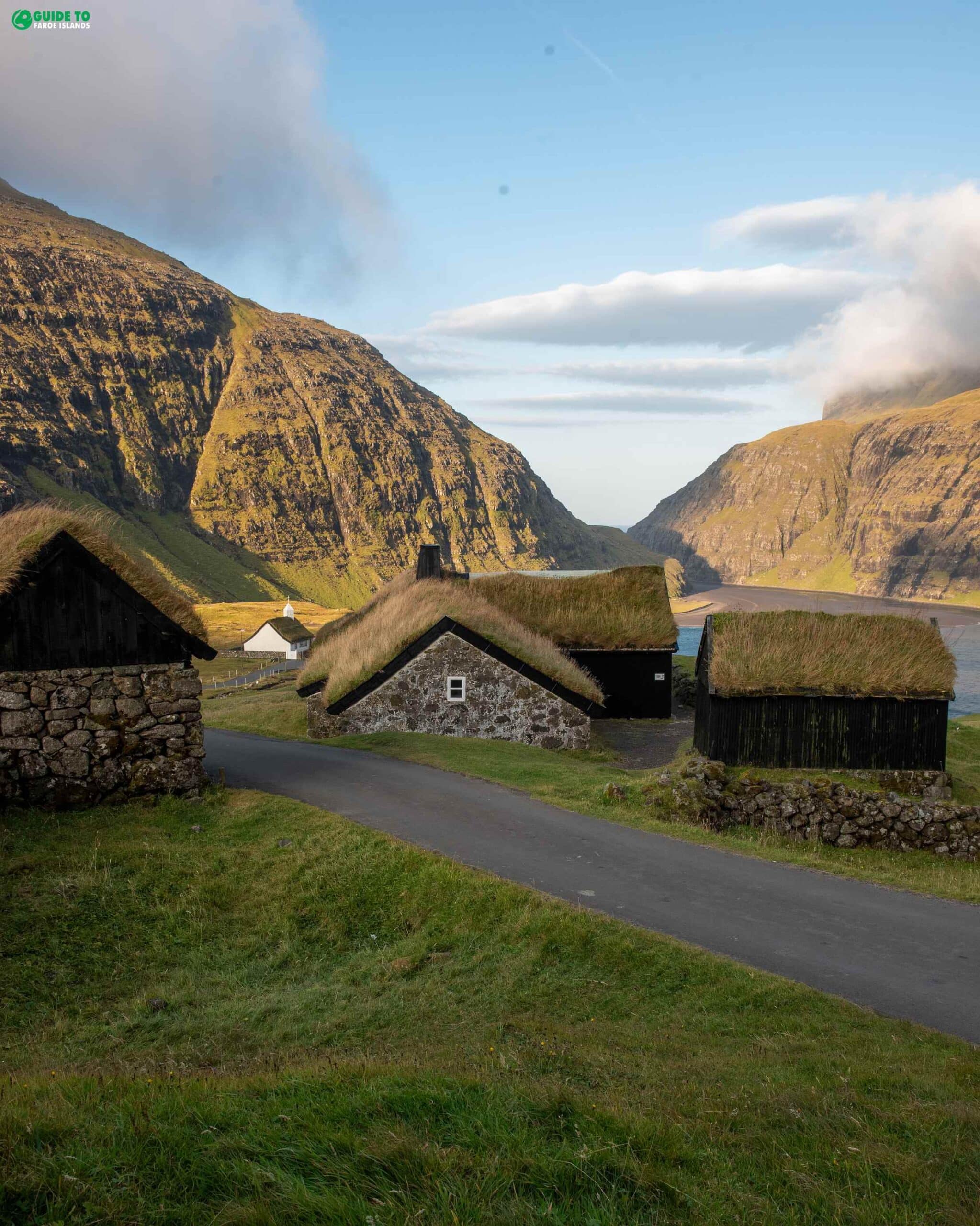 Turf-Roofed Village Houses in a Mountain Valley Black timber houses and a stone barn with grass turf roofs sit along a road in a green valley, framed by steep mountains and a distant white church near the fjord.