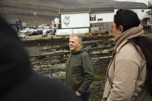Local guide sharing knowledge with visitors at the waterfront in Klaksvík, Faroe Islands, with the town's harbour buildings and hillside visible in the background