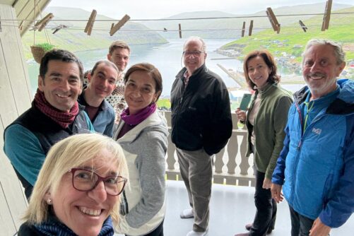 Eight people smiling for a group selfie on a covered balcony overlooking the fjord and village of Vestmanna.