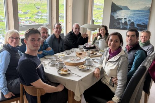 Nine people seated around a dining table for a meal, with views of the Vestmanna hillside and sea through large windows.