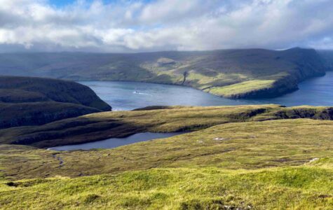 Panoramic view from the mountains north of Vestmanna on Streymoy Island, Faroe Islands, with a fjord, small mountain lake, green hillsides and a boat leaving a wake on the water below