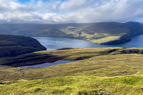 Panoramic view from the mountains north of Vestmanna on Streymoy Island, Faroe Islands, with a fjord, small mountain lake, green hillsides and a boat leaving a wake on the water below