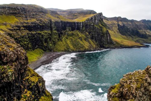 Black sand beach and turquoise water at the abandoned settlement of Víkar on Vágar Island, Faroe Islands, with towering basalt cliffs and a waterfall behind