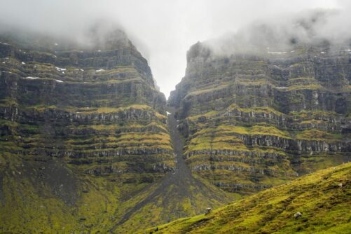 Towering layered basalt cliffs with a deep ravine at Skarð, Kunoy Island, Faroe Islands, with fog, scree slope, and sheep grazing below
