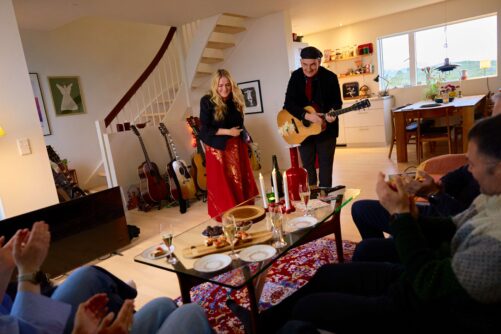 A man plays acoustic guitar while a woman in a red skirt smiles beside him during an intimate living room concert for seated guests.